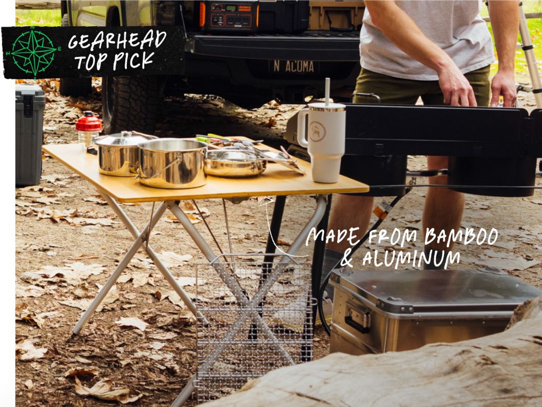 A person prepares a meal on a camp stove beside a camp table and cooler in a campsite. Text overlay reads: Gearhead Top Pick, made from bamboo & aluminum.